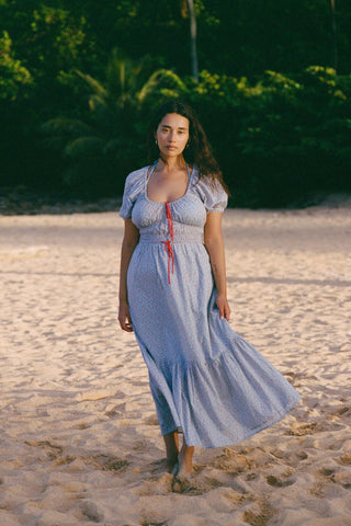 Woman in a light blue and red floral dress dress standing on a sandy beach with greenery in the background