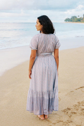 Woman in a long, light-blue colored dress standing on a beach with ocean and sky in the background