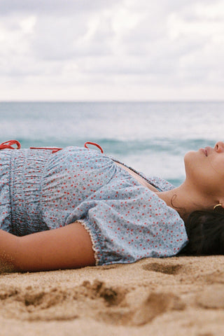 Woman in a blue and red floral dress lying on a sandy beach with ocean in the background