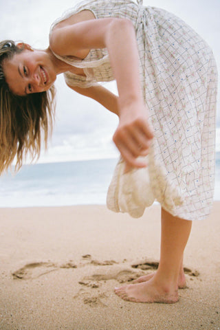 Woman in a white patterned dress standing on a beach, smiling and looking at the camera.