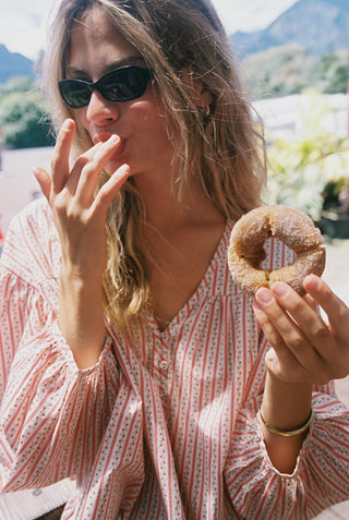 Woman in a red and white patterned shirt eating a donut with mountains in the background. 