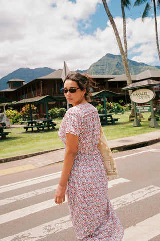 Woman looking over her shoulder at the camera, with a V-neck dress featuring a small blue and red floral pattern dress. The dress is just above ankle length. She has a woven bag over her shoulder. 