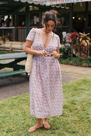 Woman holding sunglasses, with a V-neck dress featuring a small blue and red floral pattern dress. The dress is just above ankle length. She has a woven bag over her shoulder. 