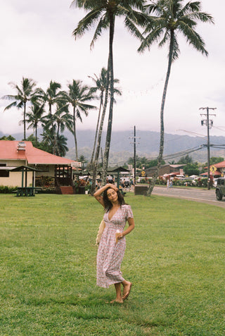 Woman standing in open grass, with a V-neck dress featuring a small blue and red floral pattern dress. The dress is just above ankle length. She has a woven bag over her shoulder. 
