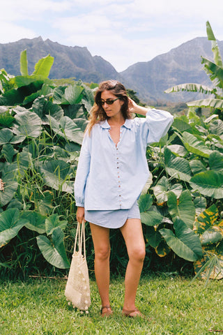 Woman in light blue chambray button-down shirt and a skort standing in a lush green field with mountains in the background