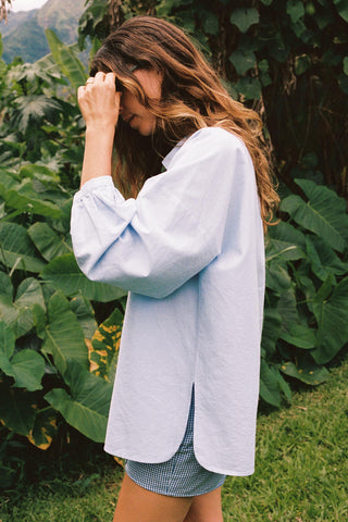 Woman wearing a light blue chambray button-down in a natural setting with green foliage.