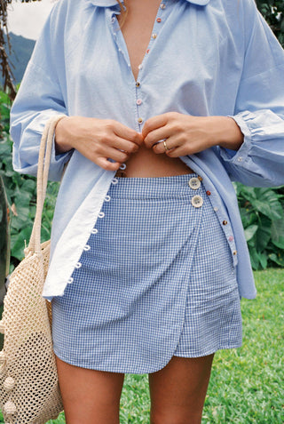 Person wearing a light blue shirt and checkered skort with a woven bag outdoors.