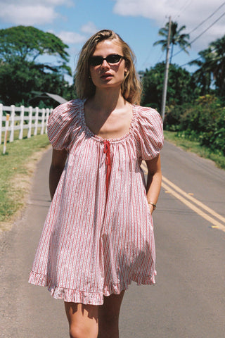 Woman in a red and white striped pattern dress standing on a road with greenery and a white fence in the background