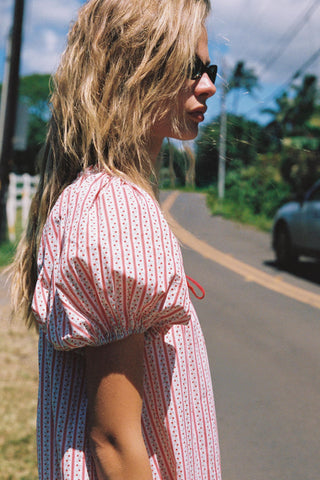 Woman in a red and white striped pattern dress standing on a road with trees and a car in the background
