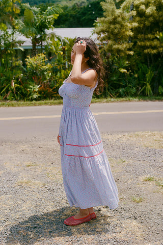 Woman in a light blue floral dress with red trim long dress standing on a road with greenery in the background