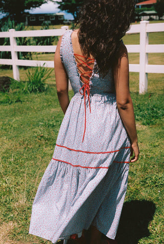 Woman in a blue dress with red trim standing in a grassy field with a white fence in the background.