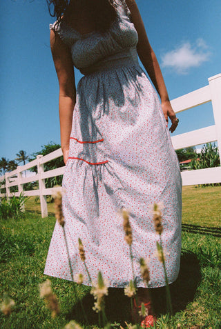 Person wearing a light blue floral dress with red trim standing in a grassy field with a white fence and blue sky in the background.