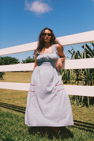 Woman in a light blue and red floral dress with red trim standing in a grassy area with a white fence and trees in the background.