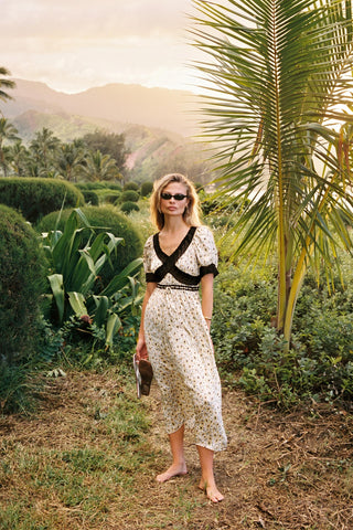 Woman in a floral dress standing in a lush garden with palm trees and mountains in the background.