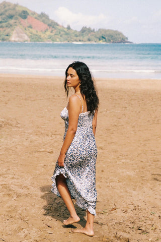 Woman in a floral dress standing on a sandy beach with ocean and mountains in the background