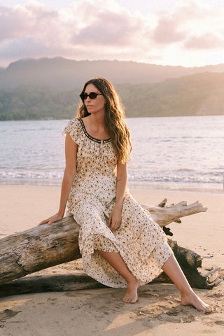 Woman in a floral dress sitting on a log by the ocean at sunset