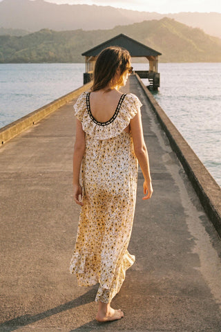 Woman in a floral dress walking along a pier by a body of water with mountains in the background.