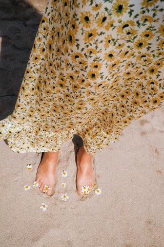 Person wearing a floral dress standing on sand with small flowers around their feet