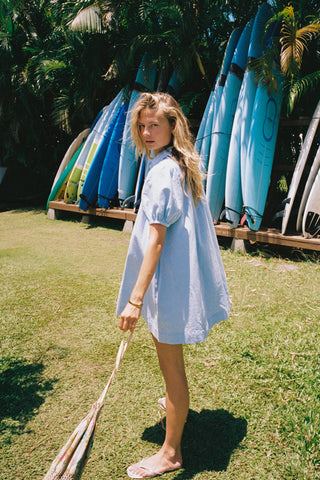 Woman in a chambray button down dress holding a paddle on grass with surfboards in the background