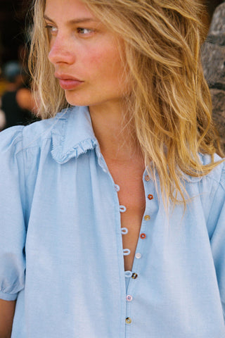 Woman in a chambray button down dress  with buttons undone, standing against a blurred background.
