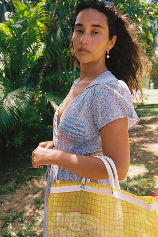 Woman in a light blue floral patterned dress holding a yellow checkered bag in a park-like setting 