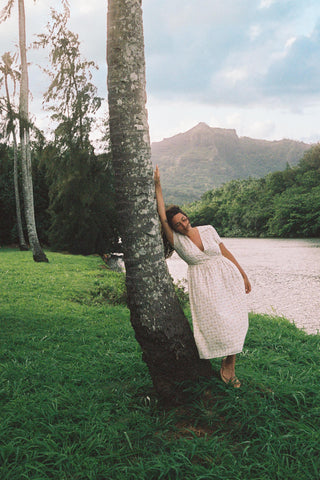 Woman in a cream patterned dress  leaning against a tree by a lake with mountains in the background