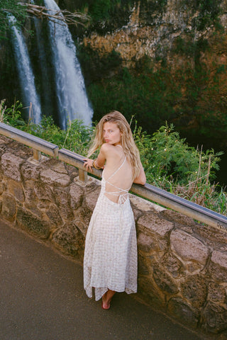 Woman in a white flowing backless dress standing by a stone wall with a waterfall in the background
