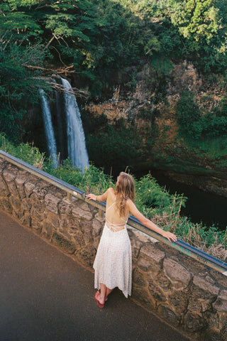 Woman in a white backless dress standing on a road looking at a waterfall in a lush green forest.