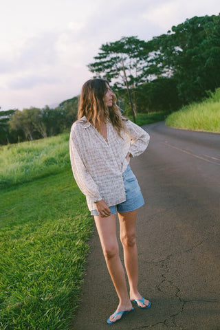 Woman wearing a checkered shirt with floral patterns standing on a road next to a grassy area with trees in the background