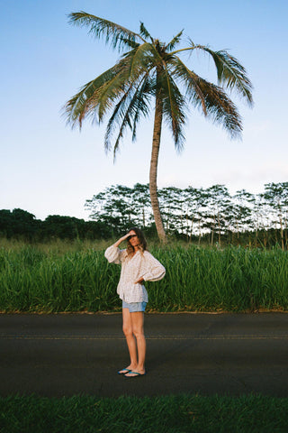 Woman wearing a checkered shirt with floral patterns standing on a road with a palm tree and greenery in the background