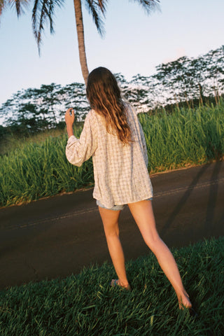 Woman wearing a checkered shirt with floral patterns walking on a path with greenery and palm trees in the background