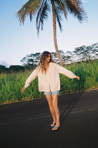 Woman wearing a checkered shirt with floral patterns standing on a road with palm trees and greenery in the background
