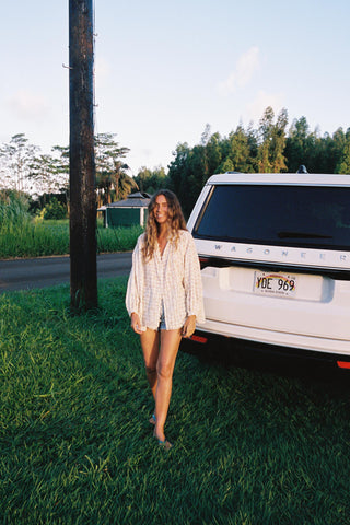 Woman wearing a checkered shirt with floral patterns standing next to a white van in a grassy area with trees in the background