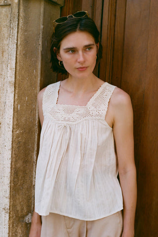 Woman wearing a light-colored sleeveless top with lace details, standing against a wooden wall.
