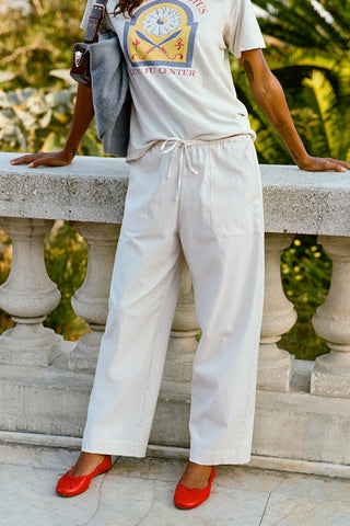 Person wearing a white t-shirt with a graphic design, white pants, and red shoes, leaning against a wall outdoors with greenery in the background.