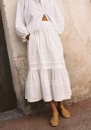 Person wearing a white outfit with a long skirt against a textured wall.