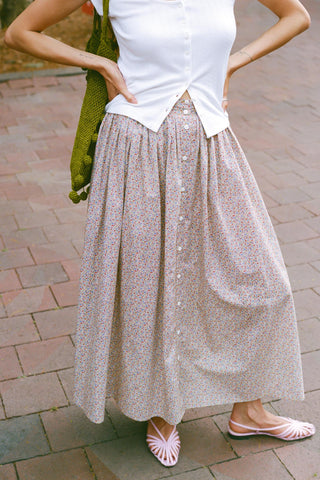 A woman wearing a long, button-front midi skirt with a small beige and pink floral print, paired with a white top and pink sandals, standing on a brick patio.