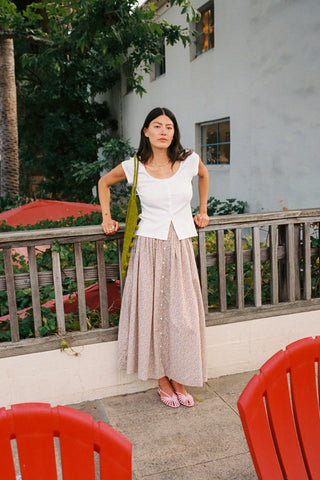 A woman stands on an outdoor patio wearing a white sleeveless top and a long, light pink patterned skirt, holding a green shoulder bag. Red chairs are visible in the foreground, with a white building and greenery in the background.