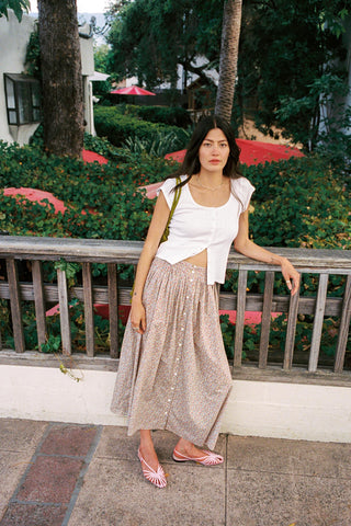 A woman stands outdoors on a tiled walkway beside a wooden railing, wearing a white sleeveless crop top and a loose, sparkly champagne-gold maxi skirt with pink sandals. Greenery and patio umbrellas are visible in the background.