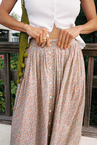 A person wearing a white cropped button-front top and a loose, high-waisted pleated skirt with a small multicolor floral print. The photo is cropped from the chest down, showing hands at the waistband and a green knit bag hanging at the side, with a wooden railing and greenery in the background.