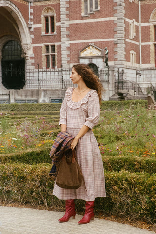 A woman stands outdoors in front of a large brick building and manicured garden hedges, wearing a long ruffled-sleeve dress in a light pink and white gingham pattern, holding a brown tote bag and a plaid jacket. She is wearing red ankle boots.