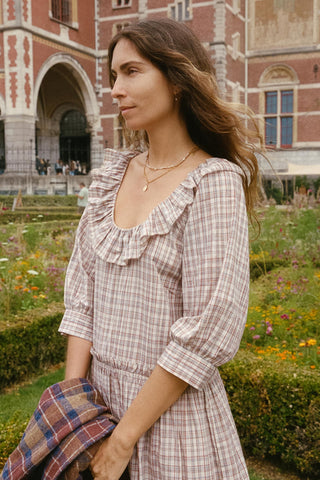 A woman wearing a beige and brown plaid dress with ruffled trim stands in a garden in front of a historic brick building. She is holding a matching plaid garment.