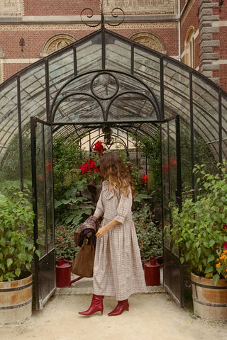 A woman in a beige plaid dress and red boots stands inside a greenhouse, holding a brown leather handbag. The greenhouse has a black metal frame with glass panels, surrounded by potted plants and red watering cans, with a brick building in the background.