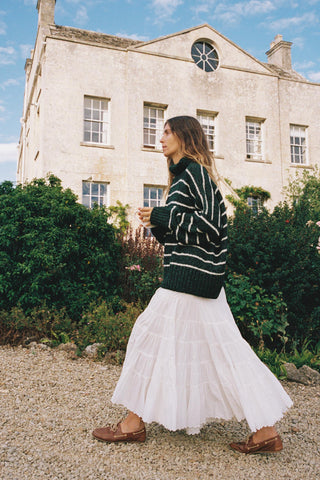 Woman in a green sweater and white skirt standing in front of a stone building with greenery.