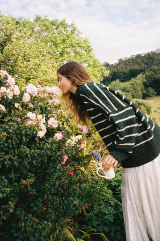 Woman in a striped sweater and white skirt standing next to a flowering bush outdoors.