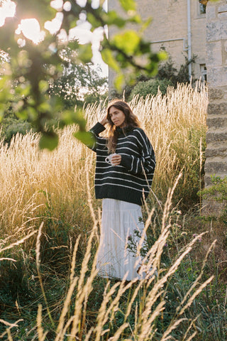 Woman in a striped sweater and white skirt standing in tall grass with a building in the background