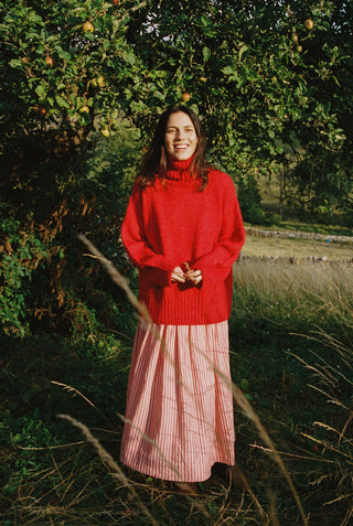 Woman in a red turtleneck sweater and pink skirt standing in a natural setting with trees and grass.