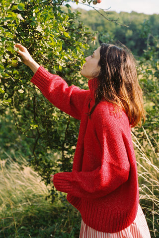 Woman in a red sweater standing in a natural setting, reaching out to touch a tree.