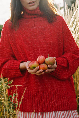 Person wearing a red sweater with a turtleneck holding apples in a field