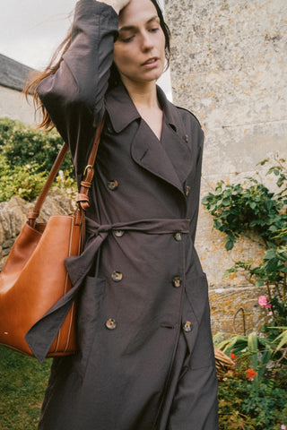 Woman wearing a dark trench coat holding a brown leather bag outdoors.